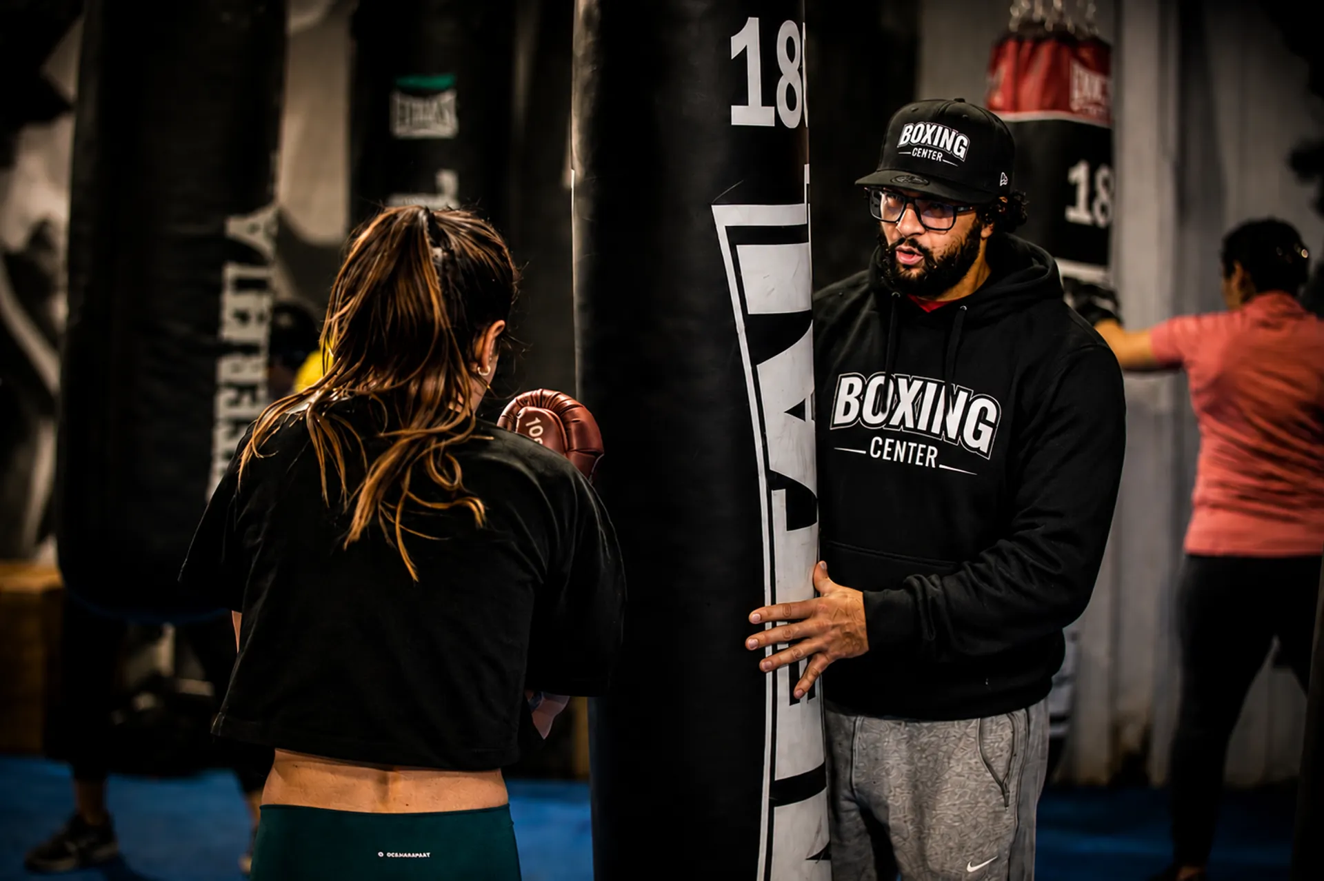 Un coach en casquette et sweat Boxing Center tient le sac de frappe pendant qu'une pratiquante travaille ses coups