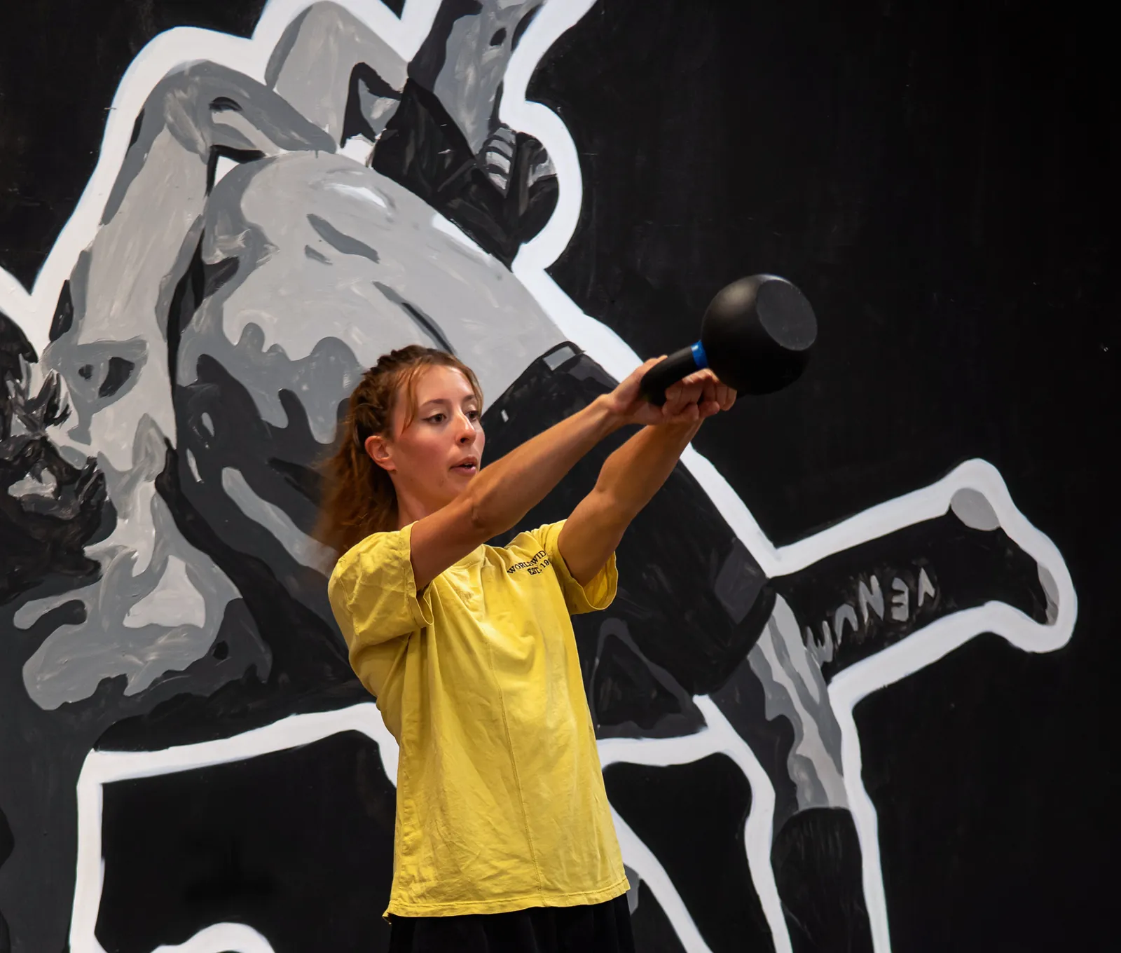 Une pratiquante en t-shirt jaune lève un kettlebell à bout de bras devant une fresque noire et blanche