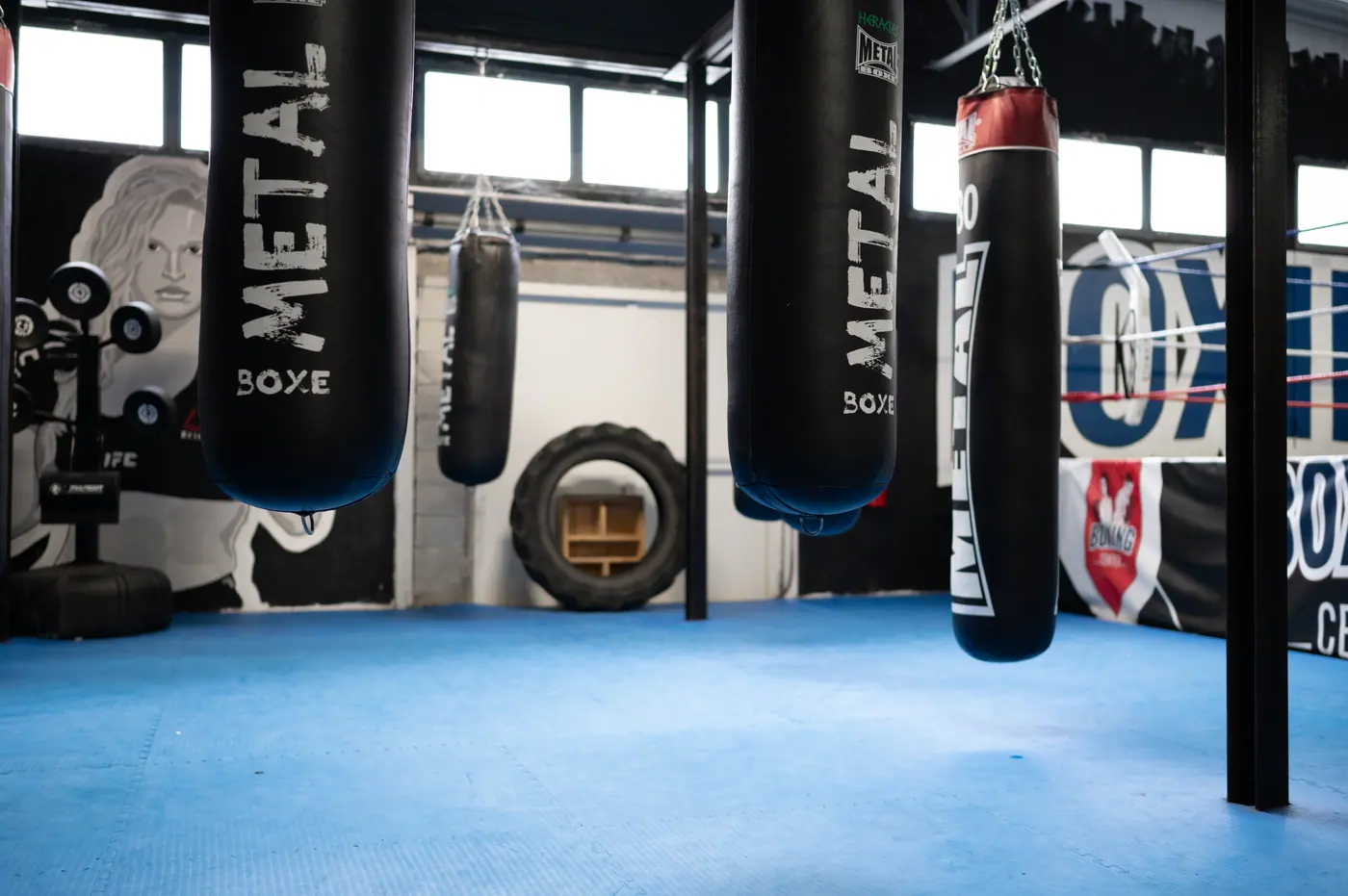 Des sacs de frappe Metal Boxe suspendus au-dessus du tapis bleu, une fresque de boxeuse au mur et la bâche du ring à droite
